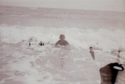 A lone figure is submerged in churning waves, appearing to struggle against the current. Black and white photograph, likely a...