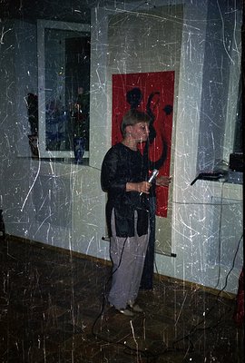A man stands at a microphone, speaking in a room with a large abstract artwork on the wall. He wears a dark suit jacket, ligh...