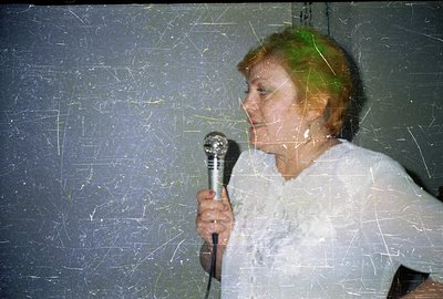 A woman holds a microphone, seemingly addressing an audience, depicted in profile. The image exhibits characteristic film gra...