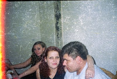 A close-up snapshot captures three people in what appears to be a dimly lit interior. A man in a light-colored shirt stands f...