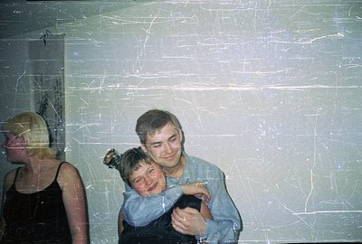 A young man in a blue shirt warmly embraces a child with dark hair and a bow, set against a textured wall. A blonde woman in ...