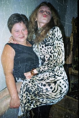 Two young women stand close, one in a simple black dress, the other in a patterned midi dress. Visible film grain & scratches...
