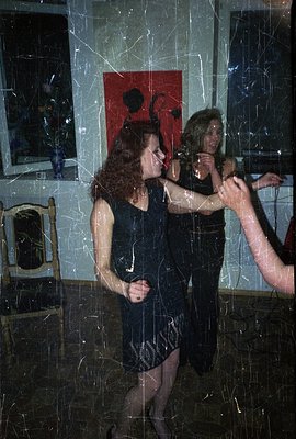 Two women dance closely, reflected in a large, scratched mirror. They wear short, sequined dresses and have striking red and ...