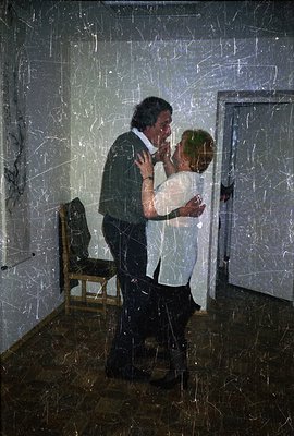 A man in a suit embraces a woman in a white shirt, standing in a sparsely furnished room. The image, likely a photograph, exh...