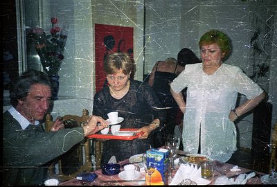 A snapshot capturing a candid moment indoors. A man in a suit is seated at a table laden with food, while two women appear to...