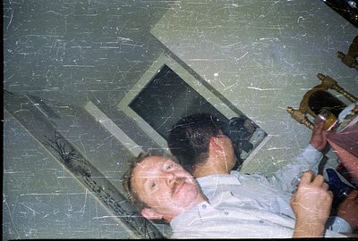 A man reclines on a floor, likely within a home, near a window and a wooden side table holding a lamp. The photograph shows v...