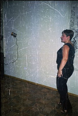 A woman in a dark, sleeveless dress stands in profile against a textured, white wall. A dark object (likely an amplifier or s...