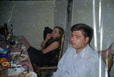 A table setting with plates of food is visible, alongside two people. A woman in a black dress sits, looking away, while a ma...