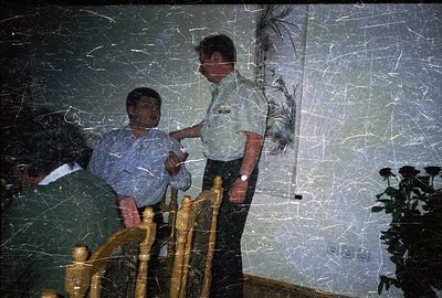 Two men interact indoors, one seated, the other standing near a railing. The scene suggests a candid moment, possibly a discu...