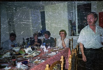 A family gathers for a dinner, seated around a table covered with a pink tablecloth. The setting appears to be a home dining ...