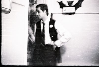 Man in a dark suit and tie holds a document or sheet music, appearing preoccupied. Strong shadow cast on wall behind, featuri...