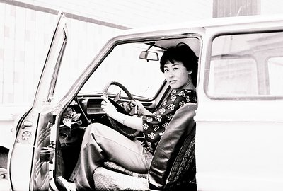A young woman sits in the driver's seat of a vintage car, likely a station wagon. She wears a patterned, dark-colored top and...