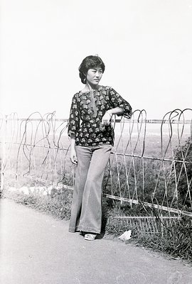 A woman poses, leaning against a barbed-wire fence. She wears wide-legged trousers and a printed, long-sleeved blouse – indic...