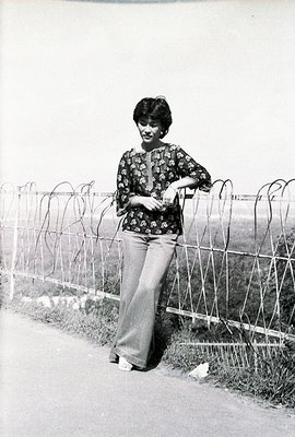 A woman in a patterned blouse and bell-bottom trousers stands near a barbed-wire fence. Mid-1970s style, likely captured in E...