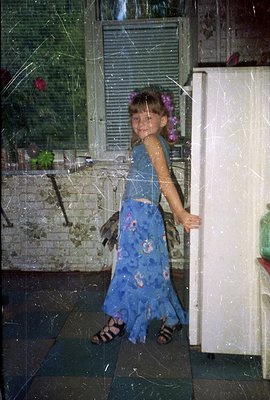 A young girl in a blue floral-print dress and sandals stands near a white refrigerator, playfully posing. The room features b...
