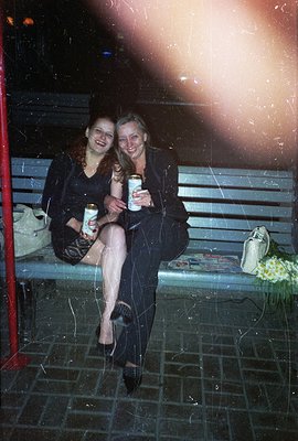 Two women, likely friends, share a moment on a park bench, each holding a beer. One wears a red dress, the other a striped su...