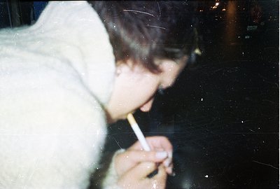 Close-up, profile view of a person in a white sweater, smoking a cigarette. Dark background suggests a nighttime setting, pot...