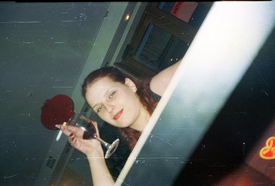 A young woman holds a wine glass in a reflective, mirrored setting. Red roses are visible in the glass. Visible grain and col...