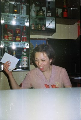 A woman in a light pink floral blouse appears to be enthusiastically reacting to something while holding a piece of paper. Ba...