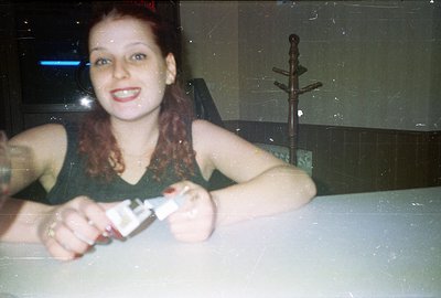 A young woman with red hair holds a bottle of alcohol, smiling directly at the camera. Interior setting with dark walls, a ne...