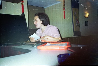 A woman with a short, dark haircut holds a paper at a table. The setting appears to be a restaurant with Asian decorative ele...