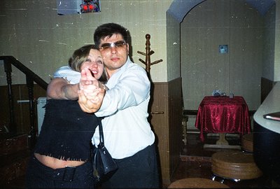 A young couple dances closely, captured in a candid moment. The man wears a white shirt & dark trousers, sporting distinctive...