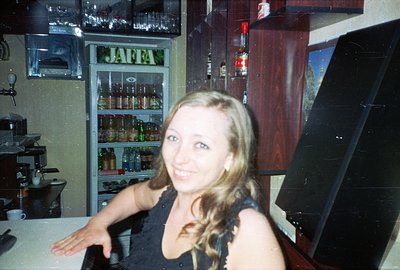 A young woman sits behind a bar, smiling at the camera. Visible are shelves filled with bottled beverages & dark wood panelin...
