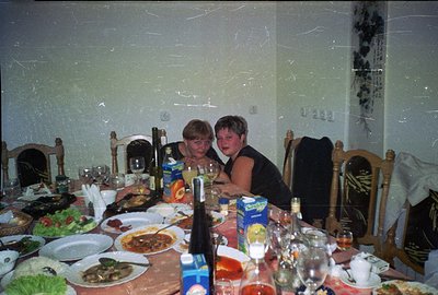 Two women sit at a table laden with food, likely enjoying a shared meal. The scene evokes a casual, informal gathering. Visib...
