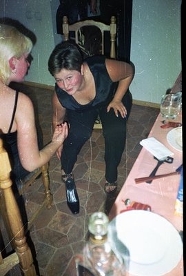 A young woman in a dark, silky top and trousers playfully poses, seated on a table. A partially visible blonde woman is acros...