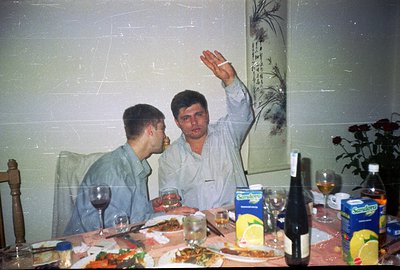 A close-up of two men at a dining table laden with food and drinks, likely during a family gathering. One raises a glass in a...