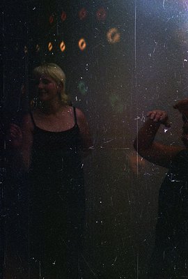 A woman in a dark dress stands near a window, blurred lights visible through the glass. Appears to be a candid moment, possib...