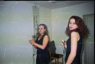 Two women, one blonde and one redhead, pose with vintage microphones in what appears to be a sparsely decorated room with tex...