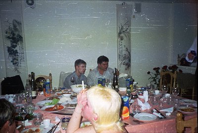 A crowded indoor scene depicting a formal dinner. Several men are seated around a long table set with glassware, plates of fo...