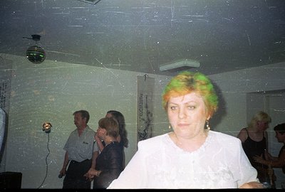 A candid snapshot captures a casual gathering, likely a party or informal event. A woman with vibrant green hair is centrally...