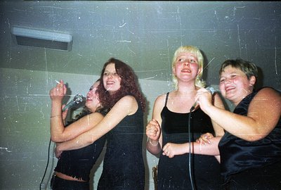 Three young women singing into microphones; cropped, black tank tops. Likely amateur snapshot, possibly a karaoke event or ca...