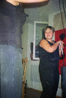 A young woman with short hair holds a microphone, seemingly on stage, next to a man partially visible on the left. The settin...