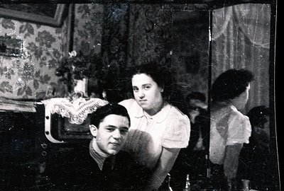 A posed studio portrait shows a young couple and a child reflected in an ornate mirror. The man, in a suit, looks directly at...