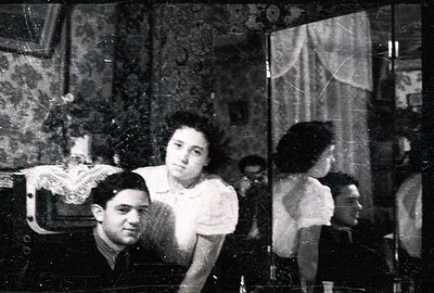 A young couple poses formally near a mirror. The woman wears a light-colored dress with puff sleeves and the man a dark suit ...
