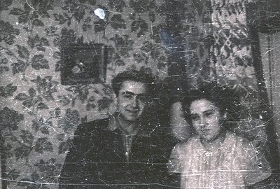 A black and white portrait shows a formally dressed man and woman. The man, with a neatly styled haircut, stands beside the w...