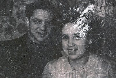 A young couple poses for a formal portrait, likely in the mid-20th century. The man, in a suit, stands slightly behind the wo...