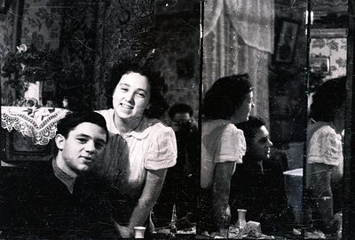 A young couple poses near a vintage mirror, likely in a domestic setting. The man wears a suit; the woman, a light-colored to...