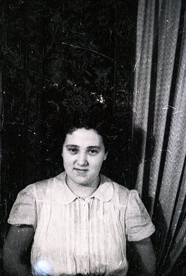 A portrait of a woman seated, wearing a collared shirt. She exhibits a direct gaze towards the camera. The background consist...