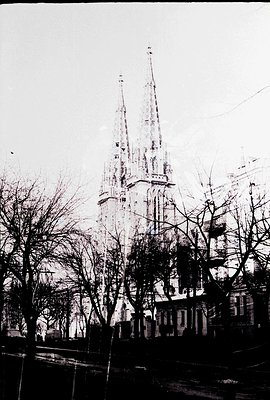 Dramatic, grayscale image of a Gothic cathedral with soaring twin spires dominating the skyline. Bare trees frame the view. L...