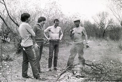 Four men stand in a wooded area observing a small fire. Three wear sweaters and striped shirts, while one is bare-chested and...