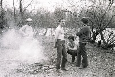Four men gathered outdoors near a smoking bonfire; one man is nude, wearing a beret. Visible are rustic foliage, a bank, and ...