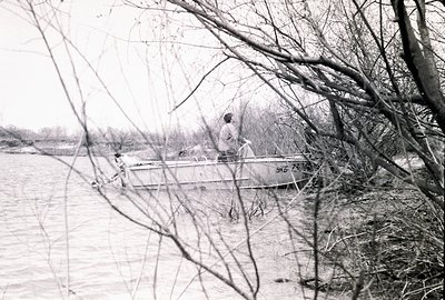 A small motorboat navigates a calm river, obscured by bare tree branches in the foreground. A single figure is visible in the...