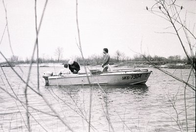 A small aluminum boat with a visible engine number "УКБ-7304" traverses a river, carrying three individuals. Two men are seat...