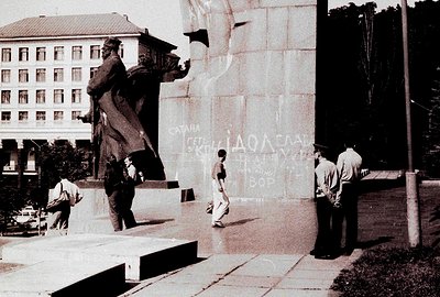 Large, imposing bronze statue of a man stands atop a concrete pedestal, viewed against a building with numerous windows. Thre...