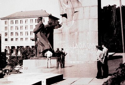 Monumental bronze statue depicting a figure holding a flag, set against a stark, institutional building. Several people are v...