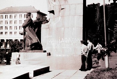 Monumental bronze statue of a soldier holding a flag, set against a stark, tall building facade. Graffiti covers the wall's s...